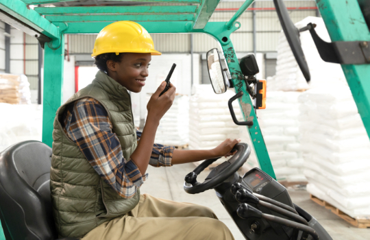 Une travailleuse conduisant un chariot élévateur à fourches dans un entrepôt se sert d'un walkie-talkie.