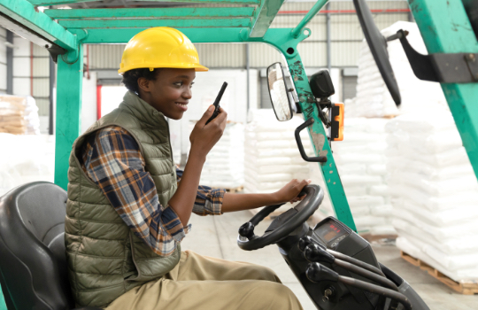 Female worker talking on walkie-talkie while driving forklift in warehouse