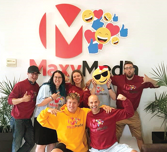 A group of Maxy Media employees standing in front of the Maxy Media logo and  smiling for the camera