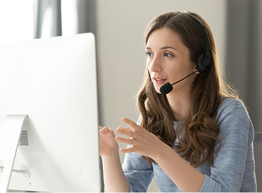 A woman facing a computer monitor speaks into a headset and gestures with her hands.