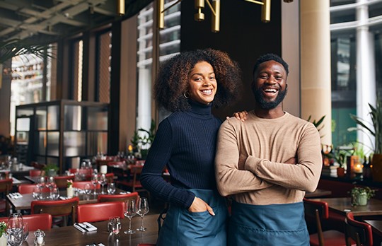 A man and woman are standing in a restaurant dining room smiling